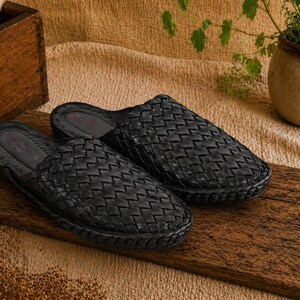 May include: A pair of black woven leather slippers. The open-back slip-on shoes are displayed on a wooden plank. The slippers have a textured, basket-weave design. The background includes a wooden box, a plant in a pot, and a tan cloth.