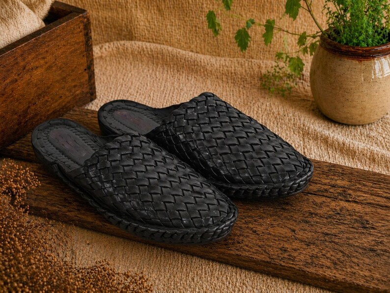 May include: A pair of black woven leather slippers. The open-back slip-on shoes are displayed on a wooden plank. The slippers have a textured, basket-weave design. The background includes a wooden box, a plant in a pot, and a tan cloth.