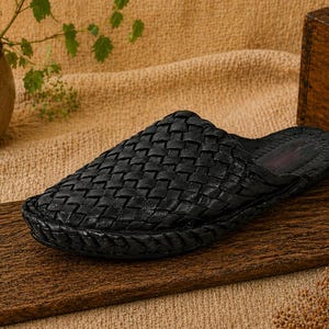 May include: A black woven leather slipper sits on a wooden surface. The slipper has a textured, basket-weave design. The background includes a small potted plant, a wooden box with the word "ORIGIN" and a tan fabric.