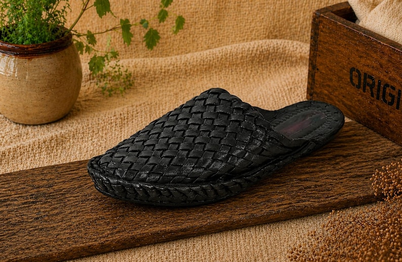 May include: A black woven leather slipper sits on a wooden surface. The slipper has a textured, basket-weave design. The background includes a small potted plant, a wooden box with the word "ORIGIN" and a tan fabric.