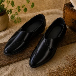 May include: A pair of black leather slip-on shoes. The shoes have a pointed toe design and a low heel. They are displayed on a wooden surface with a neutral-colored background. A small potted plant and a wooden box are also in the image.