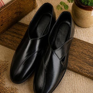 May include: A pair of black leather slip-on shoes with a glossy finish. The shoes have a unique design with a cutout detail on the side and stitched accents. They are displayed on a wooden surface.