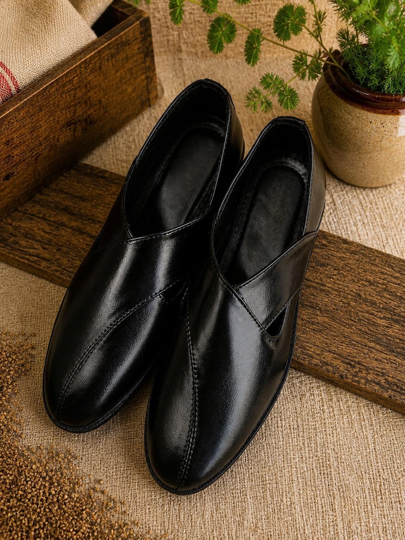 May include: A pair of black leather slip-on shoes with a glossy finish. The shoes have a unique design with a cutout detail on the side and stitched accents. They are displayed on a wooden surface.