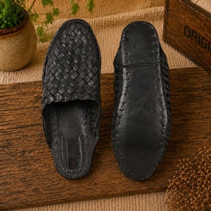 May include: Two black leather woven sandals are displayed on a wooden surface. The sandals have a slip-on design with a textured, woven upper. The soles are smooth and dark, with visible stitching. The sandals are likely handmade.