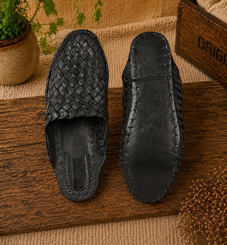 May include: Two black leather woven sandals are displayed on a wooden surface. The sandals have a slip-on design with a textured, woven upper. The soles are smooth and dark, with visible stitching. The sandals are likely handmade.