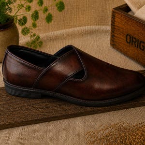 Puede incluir: Un zapato sin cordones de cuero marr&oacute;n con punta redondeada y tac&oacute;n bajo. El zapato tiene un acabado liso y pulido y una correa decorativa en la parte superior. El zapato se exhibe sobre una tabla de madera.