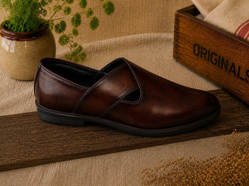 Puede incluir: Un zapato sin cordones de cuero marr&oacute;n con punta redondeada y tac&oacute;n bajo. El zapato tiene un acabado liso y pulido y una correa decorativa en la parte superior. El zapato se exhibe sobre una tabla de madera.