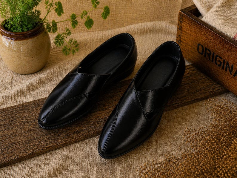May include: A pair of black leather slip-on shoes. The shoes have a pointed toe design and a low heel. They are displayed on a wooden surface with a neutral-colored background. A small potted plant and a wooden box are also in the image.
