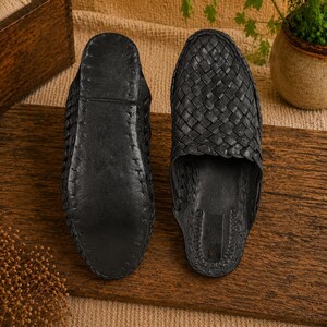 May include: Two black leather woven slippers are displayed side by side on a wooden surface. The slippers have a slip-on design with a woven upper and a solid sole. The background includes a wooden box and a small potted plant.