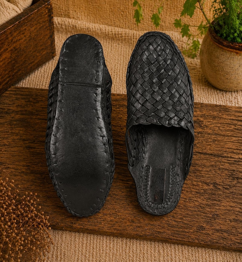 May include: Two black leather woven slippers are displayed side by side on a wooden surface. The slippers have a slip-on design with a woven upper and a solid sole. The background includes a wooden box and a small potted plant.