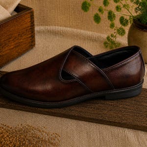 Puede incluir: Un solo mocas&iacute;n de cuero marr&oacute;n oscuro con punta redondeada y una correa en la parte superior. El zapato est&aacute; sobre una superficie de madera, con un fondo neutro. El zapato parece sin usar.