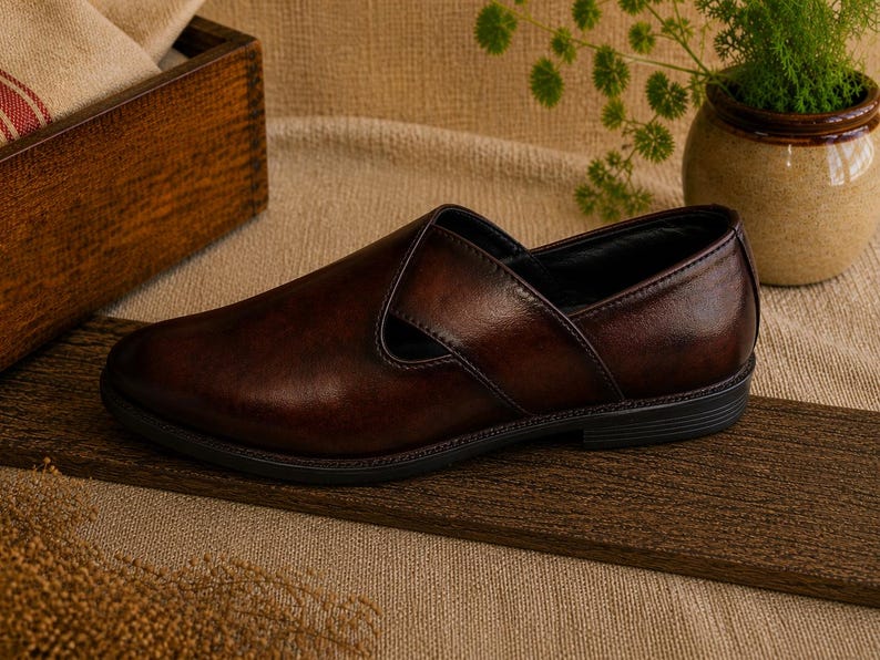 Puede incluir: Un solo mocas&iacute;n de cuero marr&oacute;n oscuro con punta redondeada y una correa en la parte superior. El zapato est&aacute; sobre una superficie de madera, con un fondo neutro. El zapato parece sin usar.