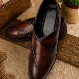 Puede incluir: Un par de zapatos sin cordones de cuero marr&oacute;n con punta en pico y un dise&ntilde;o en forma de V en la pala. Los zapatos tienen un acabado brillante y est&aacute;n sobre una superficie de madera. El fondo incluye una caja de madera y una peque&ntilde;a planta en maceta.