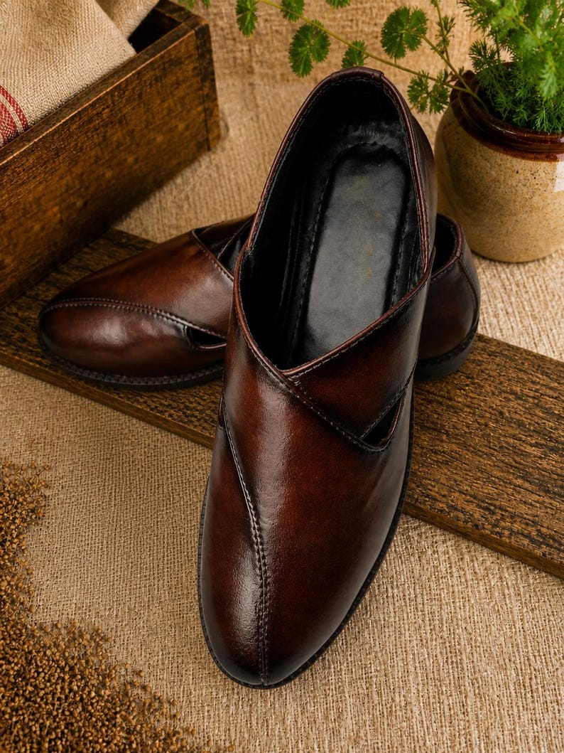 Puede incluir: Un par de zapatos sin cordones de cuero marr&oacute;n con punta en pico y un dise&ntilde;o en forma de V en la pala. Los zapatos tienen un acabado brillante y est&aacute;n sobre una superficie de madera. El fondo incluye una caja de madera y una peque&ntilde;a planta en maceta.