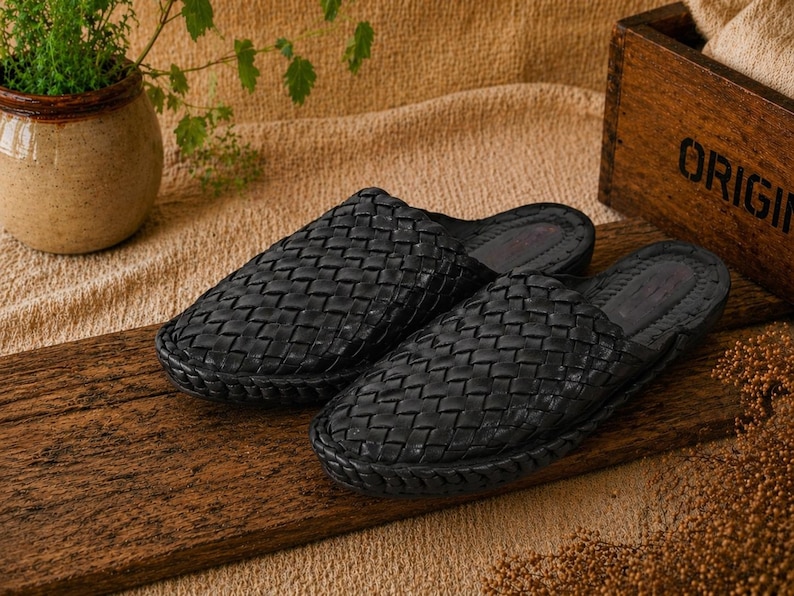 May include: A pair of black woven leather slip-on shoes. The shoes are displayed on a wooden surface, with a woven pattern across the top. The shoes are a dark black color and appear to be new.