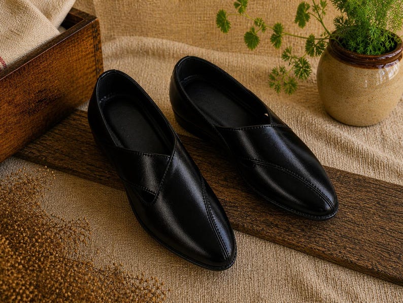 May include: A pair of black leather slip-on shoes with a pointed toe. The shoes are displayed on a wooden plank, with a neutral-toned background. The shoes have a stitched design on the top.