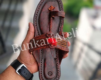 May include: A dark brown leather sandal featuring a toe post design, a wide strap across the midfoot with decorative stitching, and a small red fabric accent. The sole has a visible number one embossed near the heel, and the text BunkariAtelier.