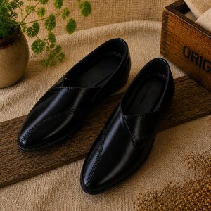 May include: A pair of black leather slip-on shoes with a pointed toe, displayed on a wooden plank. The shoes have a smooth, glossy finish. The background includes a small ceramic pot with greenery and a wooden box with the word "ORIGINALS".