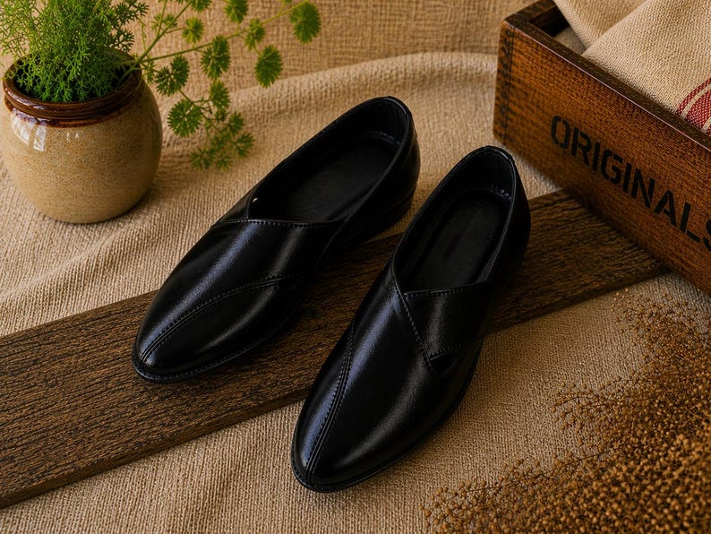 May include: A pair of black leather slip-on shoes with a pointed toe, displayed on a wooden plank. The shoes have a smooth, glossy finish. The background includes a small ceramic pot with greenery and a wooden box with the word "ORIGINALS".