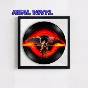 May include: Framed black vinyl record featuring a striking image of a figure with large wings against a fiery red and orange background. The text "REAL VINYL" is displayed above the record in blue.