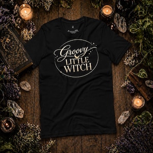 Op de afbeelding: Zwart t-shirt met de tekst "Groovy Little Witch" in een wit, cirkelvormig ontwerp. Het shirt ligt op een houten oppervlak, omringd door kaarsen, kristallen en groen, wat een mystieke uitstraling creëert.