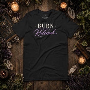 May include: Black t-shirt with the text "BURN the Rulebook" in white and purple script. The shirt is surrounded by candles, crystals, and dried flowers on a wooden surface, creating a mystical aesthetic.