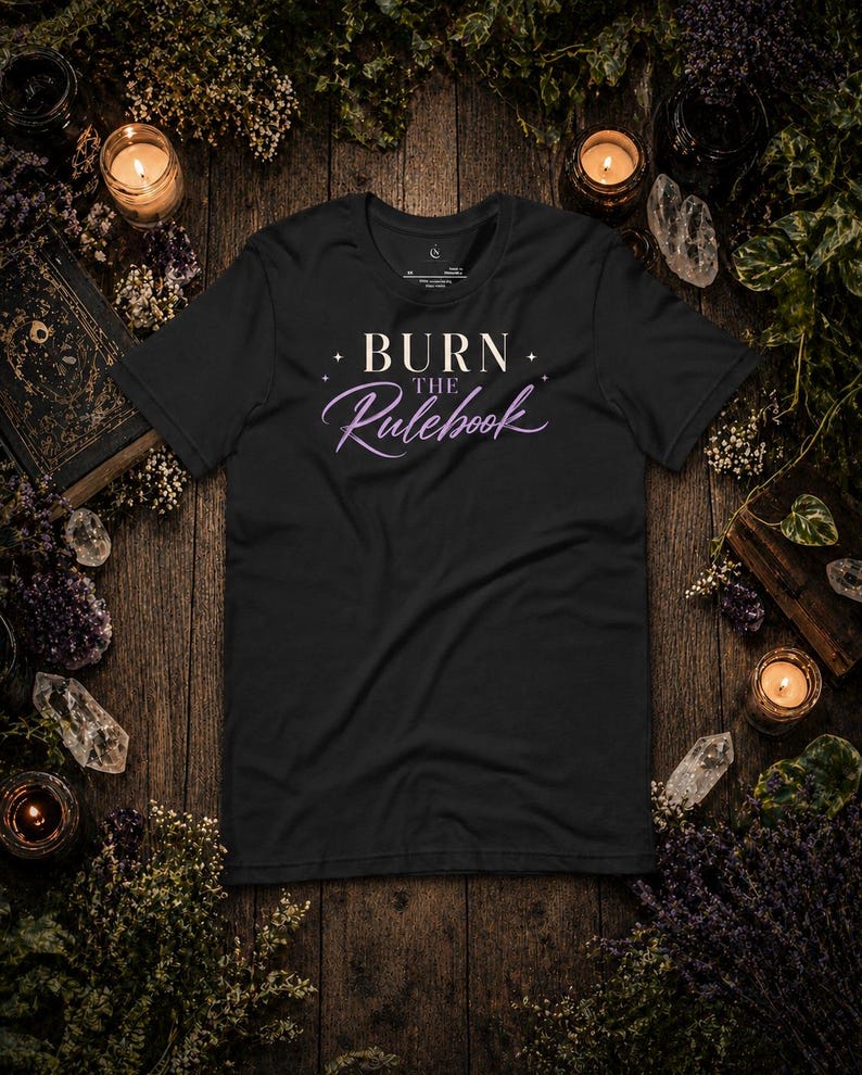 May include: Black t-shirt with the text "BURN the Rulebook" in white and purple script. The shirt is surrounded by candles, crystals, and dried flowers on a wooden surface, creating a mystical aesthetic.