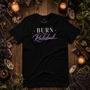 May include: Black t-shirt with the text "BURN THE Rulebook" in white and purple script. The shirt is surrounded by candles, crystals, and dried flowers on a wooden surface.