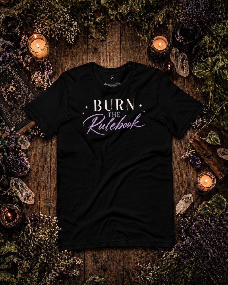 May include: Black t-shirt with the text "BURN THE Rulebook" in white and purple script. The shirt is surrounded by candles, crystals, and dried flowers on a wooden surface.