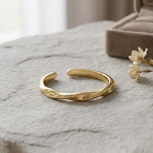 Könnte beinhalten: Ein goldfarbenes, offenes Armband mit einer glatten, wellenförmigen Oberflächenstruktur. Das Metallband hat eine polierte Oberfläche und ein durchgehendes, unregelmäßiges Wellenmuster, das für ein fließendes Erscheinungsbild sorgt.