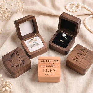 Könnte beinhalten: Drei quadratische Ringkästchen aus Holz mit Klappdeckeln. Zwei sind aus dunklem Walnussholz mit weißen oder schwarzen Samteinlagen für Ringe. Eines ist aus hellem Holz mit der Gravur ANTHONY und EDEN 06.02.2025. Andere zeigen Gravuren von Händen.