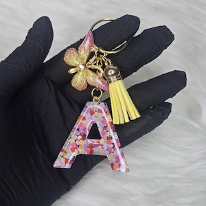 May include: A keychain featuring the letter "A" in a clear resin filled with colorful confetti. The keychain includes a gold-colored butterfly charm with pink accents, a yellow tassel, and a gold keyring.