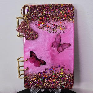 May include: A pink resin journal with two butterfly illustrations and a glittery top border. The journal has gold-colored metal rings and a heart-shaped charm attached to the side. The journal is displayed on a clear stand.