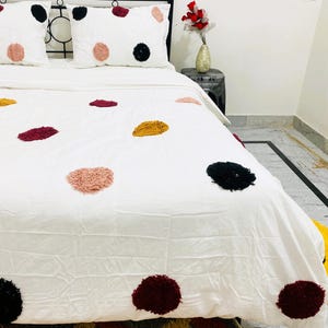 May include: White bedspread and pillow set with large, textured polka dots in black, burgundy, pink, and gold. The bedding is on a black metal bed frame. A vase of red flowers sits on a side table.