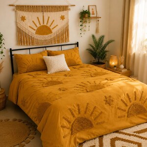 May include: A bedroom scene featuring a bed with a mustard yellow comforter and matching pillows, adorned with sunburst designs. A macrame wall hanging with a sun motif hangs above the bed. The room is decorated with plants and a jute rug.