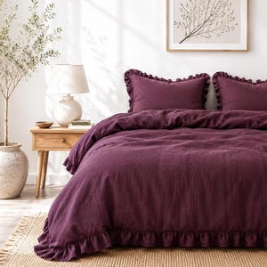 Puede incluir: Un juego de cama de color ciruela intenso que incluye una funda nórdica y dos fundas de almohada a juego, cada una adornada con bordes de volantes. La tela tiene un tejido texturizado visible y el conjunto se presenta sobre una cama con volantes.
