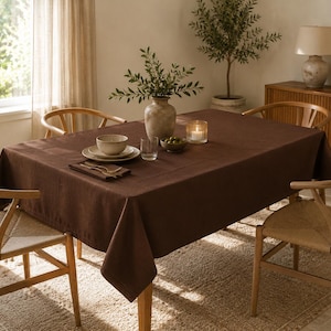 Puede incluir: Un mantel rectangular de color marrón oscuro sobre una mesa de comedor, rodeada por cuatro sillas de madera clara con asientos trenzados. La mesa está puesta con un cuenco de cerámica, platos, un vaso, un cuenco de aceitunas, una vela y un jarrón.