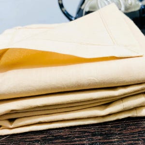 Op de afbeelding: Een stapel gevouwen, lichtgele stoffen items, mogelijk linnen of beddengoed. De items zijn netjes gerangschikt, met zichtbare stiksels langs de randen. De stof lijkt zacht en glad, wat wijst op een comfortabele textuur.