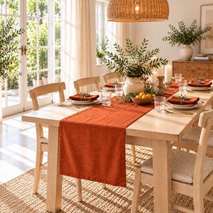 Può includere: Un lungo runner da tavola rettangolare in lino color arancio bruciato centrato su un tavolo da pranzo in legno chiaro. Il tavolo è apparecchiato con piatti bianchi, tovaglioli arancioni piegati, bicchieri trasparenti e un vaso centrale.