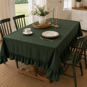 May include: A dark green linen tablecloth with a ruffled edge covers a wooden table set for a meal. The table is set with white plates, a teacup, and a gold fork. Two candlesticks with pink and green candles are on the table.