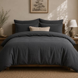 May include: A dark grey bed set with a duvet cover and pillowcases. The bed is made up with a neutral-coloured rug and wooden bedside tables on either side. A lamp and vase with greenery are visible.