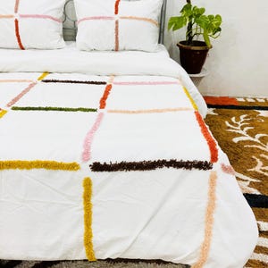 Puede incluir: Juego de cama blanco con diseño geométrico afelpado y colorido. El juego incluye una funda nórdica y dos fundas de almohada. El diseño presenta líneas que se cruzan en tonos de amarillo, naranja, rosa, marrón y verde. La cama está contra una pared blanca.