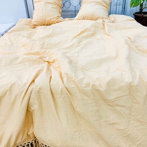 Pode incluir: Um conjunto de cama amarelo claro composto por um edredão grande e duas almofadas retangulares a condizer. O edredão apresenta uma orla com franjas decorativas na parte inferior. O tecido tem uma textura macia e ligeiramente enrugada.