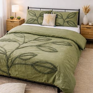Puede incluir: Una cama con un edredón verde y fundas de almohada a juego, adornada con un diseño de hojas. Una almohada decorativa con un patrón de sol se encuentra sobre la cama. La habitación cuenta con mesitas de noche de madera, una lámpara y obras de arte enmarcadas.
