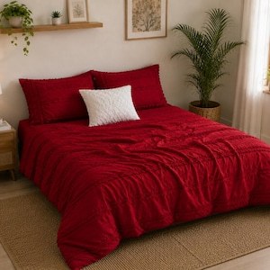 May include: A red bedspread with matching pillowcases and a white decorative pillow. The bedding is on a bed with a woven rug. A wooden bedside table with a lamp and plants is to the left of the bed. A large potted plant is in the background.