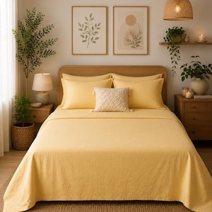 Könnte beinhalten: Ein Schlafzimmer mit einem Bett mit gelber Bettwäsche, einschließlich eines flachen Lakens, eines Spannbetttuchs und Kissenbezügen. Ein strukturiertes Kissen liegt auf den Kissen. Der Raum ist mit gerahmten botanischen Drucken, einer gewebten Pendelleuchte und Topfpflanzen dekoriert.