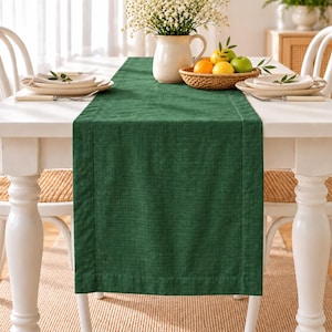 Puede incluir: Un camino de mesa rectangular de color verde oscuro con una textura tejida visible y un borde cosido. El camino está colocado sobre una mesa de comedor blanca, entre dos juegos de platos blancos y una cesta central con fruta fresca.