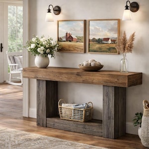 Może przedstawiać: Rustykalna drewniana konsola z naturalnym wykończeniem i szarymi filarami. Stół zdobi biały wazon z kwiatami, drewniana miska i oprawione obrazy krajobrazów. Dwa kinkiety są zamontowane nad obrazami.