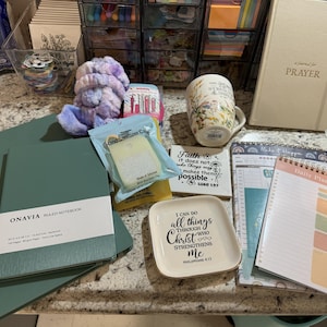 Puede incluir: Un surtido de artículos de papelería e inspiración, que incluyen un cuaderno verde, una taza floral, un diario de oración y una agenda diaria. También se incluyen un pequeño plato decorativo con una cita religiosa, un soporte para desinfectante de manos y una diadema.