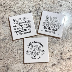 May include: Three white square coasters with inspirational quotes and floral designs. One reads "Faith it does not make things easy it makes them possible Luke 1:37." Another says "Be The Light Matthew 5:14." The third says "I will not be shaken Psalm 16:8."